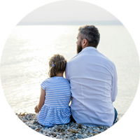 En man och en liten flicka sitter bredvid varandra på en stenig strand och tittar ut över ett lugnt, solbelyst hav under en ljus himmel.