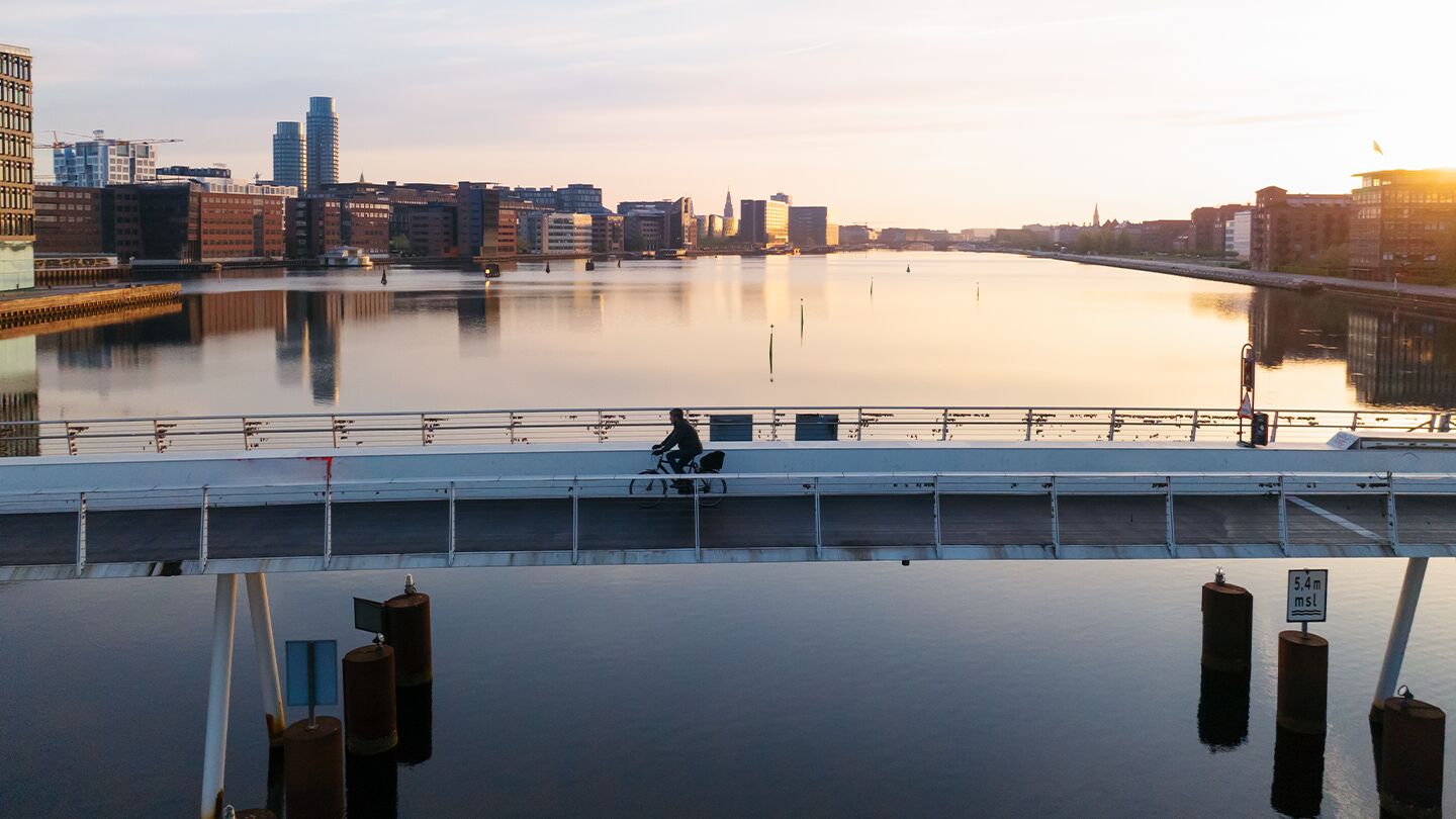 Stadssilhuett som speglas i en lugn flod med en person som cyklar på bron.