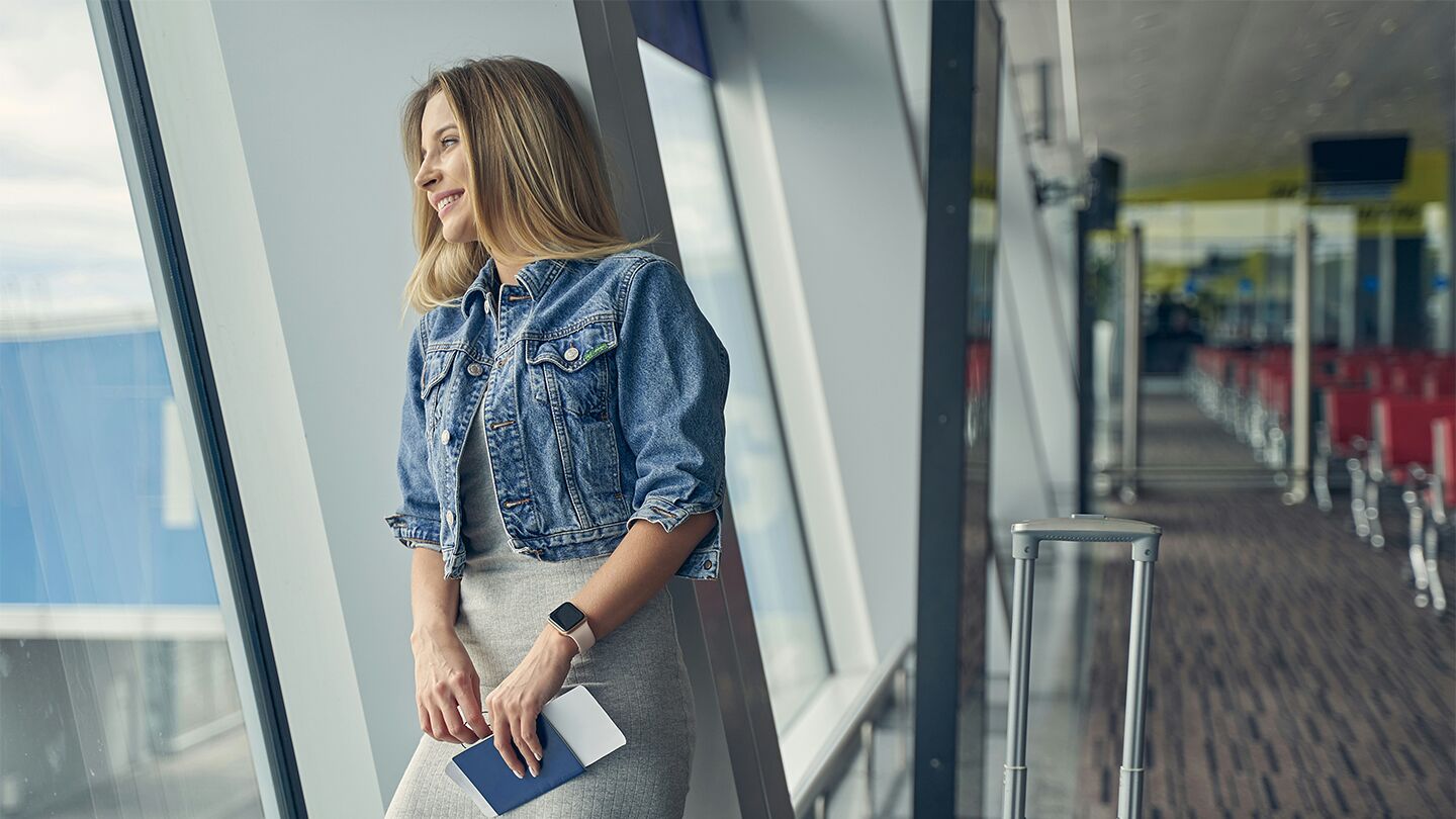 Person vid en flygplatsfönster håller i ett pass och står bredvid en kabinväska.
