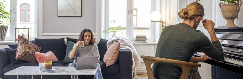 medelålders par i en lägenhet där kvinnan sitter i soffan och ler med en kaffekopp i handen och mannen sitter vid ett piano