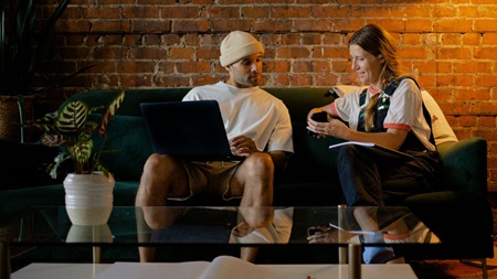 Två personer sitter på en soffa och samtalar, med en bärbar dator och anteckningsblock.