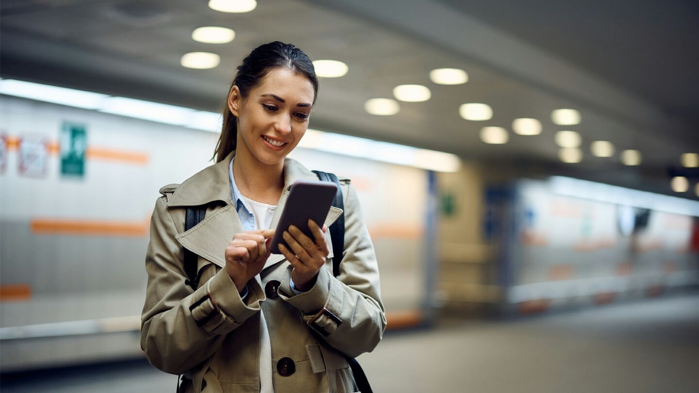 Kvinna i beige kappa står i tunnelbanemiljö och använder en mobiltelefon.