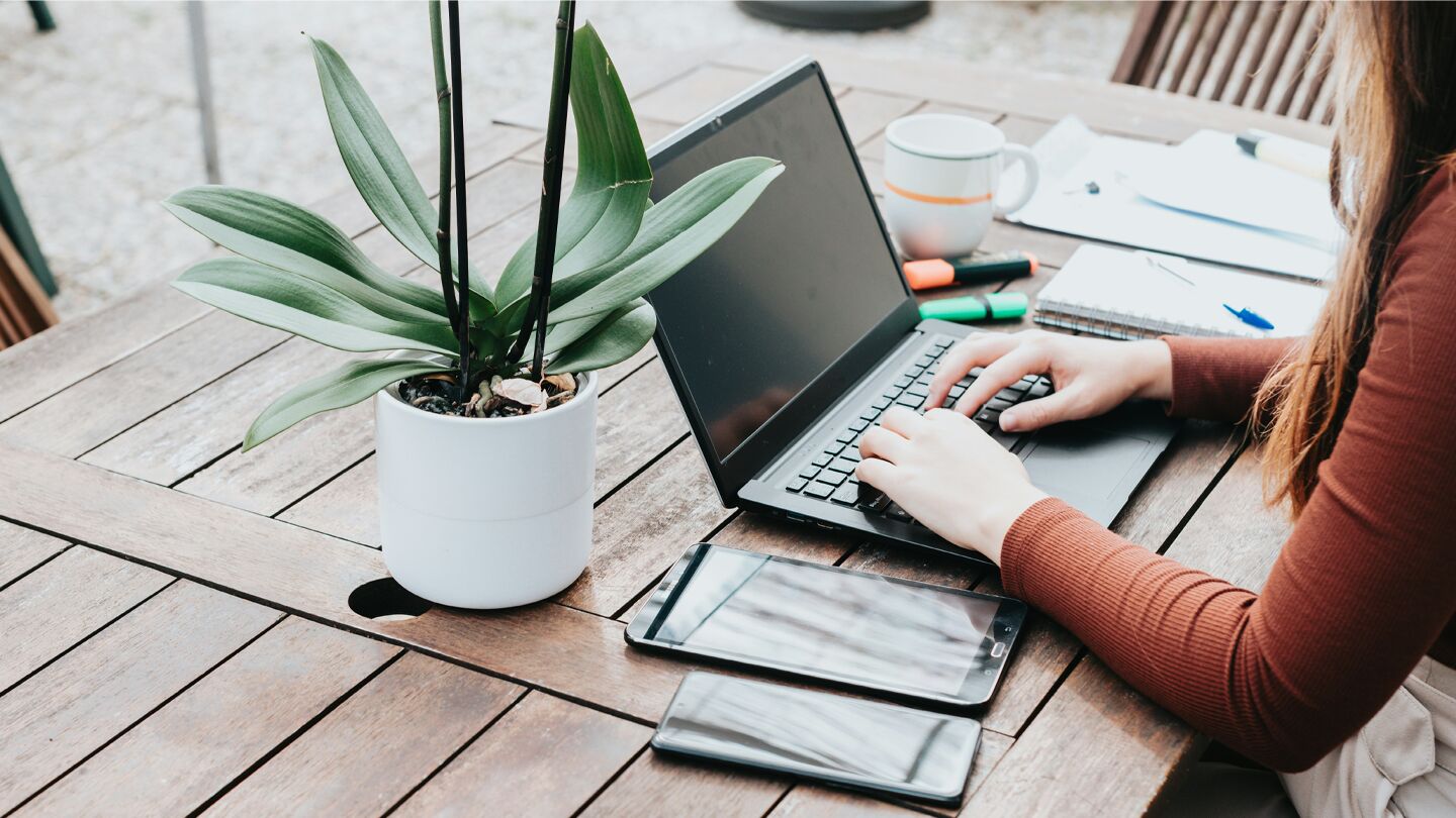 Person arbetar vid en laptop på ett träbord utomhus, omgiven av anteckningar, en kopp och elektronik.
