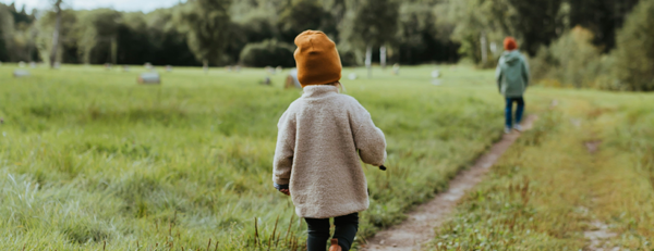 Två barn promenerar på en stig genom en grönskande äng omgiven av träd.