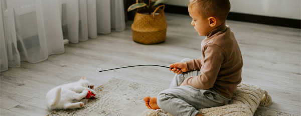 Ett barn sitter på en kudde på golvet och leker med en vit katt och en leksak. Rummet är ljust med trägolv, gardiner och en växt i en korg i bakgrunden.