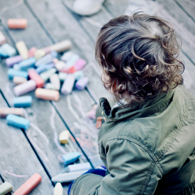 Ett barn med lockigt hår sitter på ett trädäck och tittar på färgglada kritor som ligger utspridda på marken. Barnet bär en grön jacka.