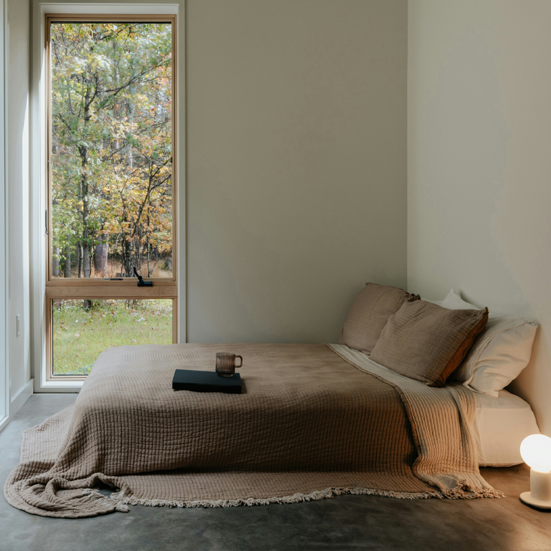 Ett minimalistiskt sovrum med en bäddad säng i jordnära toner, en lampa på golvet och ett stort fönster som vetter mot en skog.