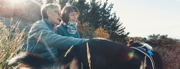 En utomhusbild med två personer och en hund, omgiven av gräs och träd med soligt bakgrund.