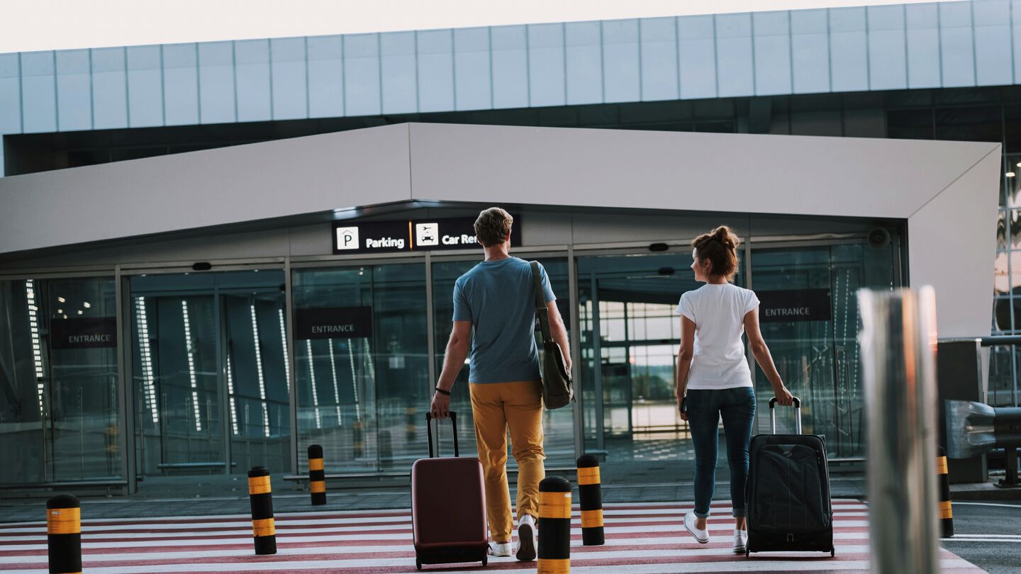 Två personer med rullväskor går mot en flygplatsterminal.
