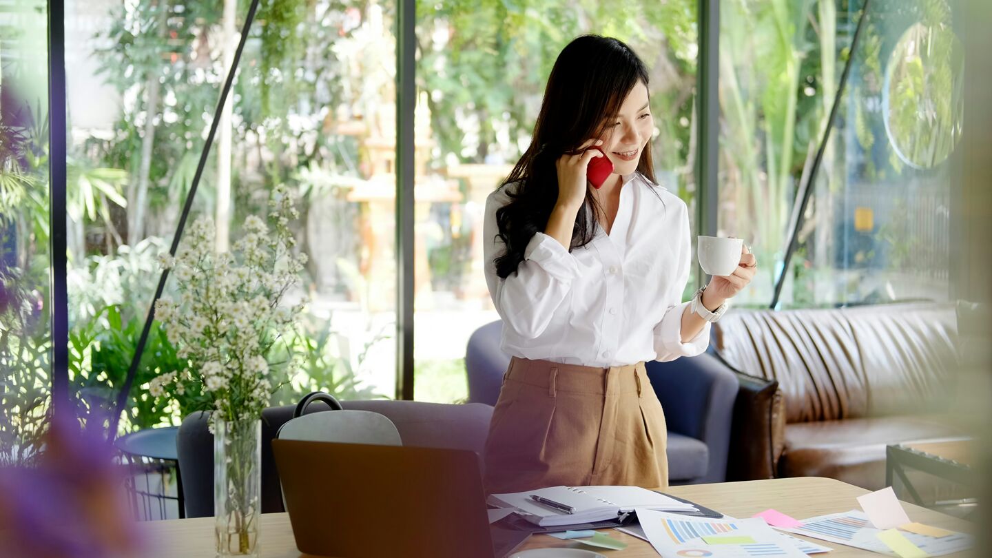 En person står vid ett skrivbord med en kopp i handen och pratar i telefon, omgiven av papper och en bärbar dator, med utsikt över en grönskande bakgrund.