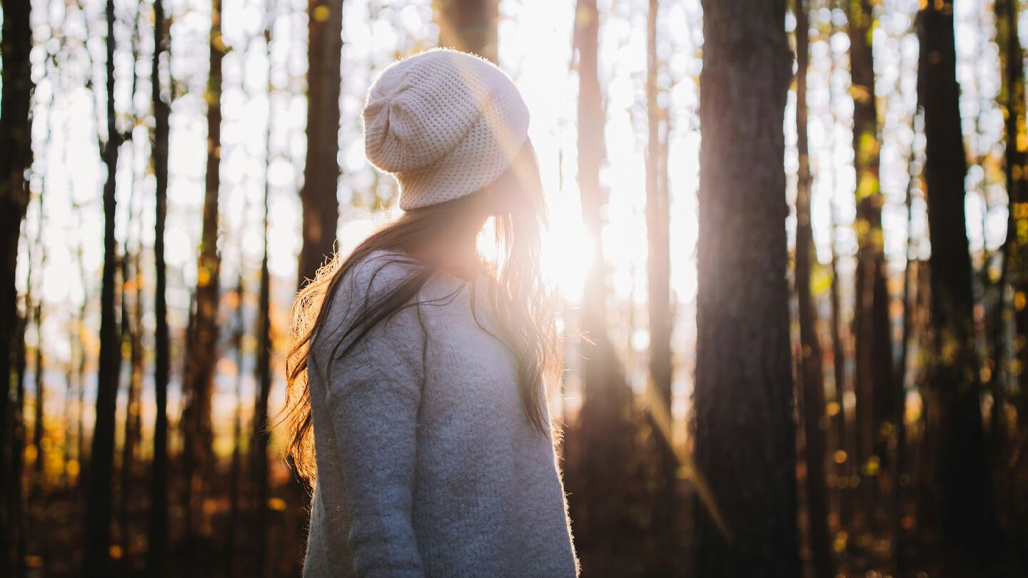En person står i en solbelyst skog, klädd i en vit mössa och ljus tröja. Solstrålarna lyser genom träden i bakgrunden.