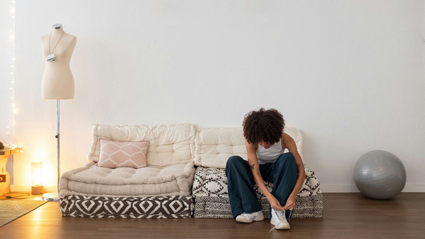 En person sitter på en låg soffa i ett ljust rum, knyter sina skor. En provdocka, en lampa och en pilatesboll syns i bakgrunden.