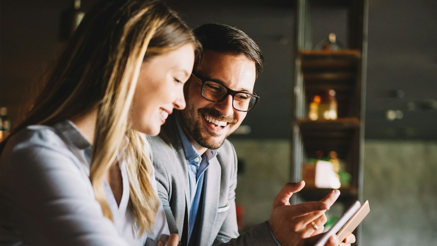 Två personer samtalar och tittar på en surfplatta i en inomhusmiljö med hyllor i bakgrunden.