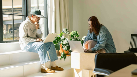 Två personer sitter i ett ljust rum och arbetar på sina bärbara datorer. Den ena personen sitter på en trappa vid ett fönster, medan den andra sitter vid ett bord. En grön växt står på bordet mellan dem, och solljus strömmar in genom fönstret och lyser upp rummet.
