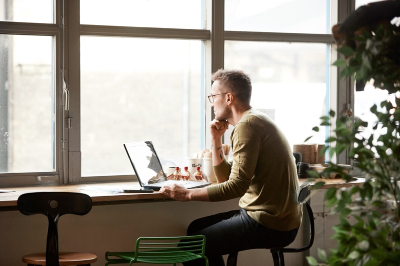 En person sitter vid ett fönster och arbetar på en bärbar dator, omgiven av växter och föremål på ett skrivbord i en ljus miljö.
