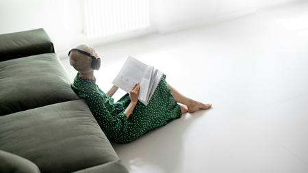 En person sitter på golvet intill en grön soffa, iklädd en grön klänning med vita prickar, lyssnar på hörlurar och läser en bok i ett ljust, minimalistiskt rum.