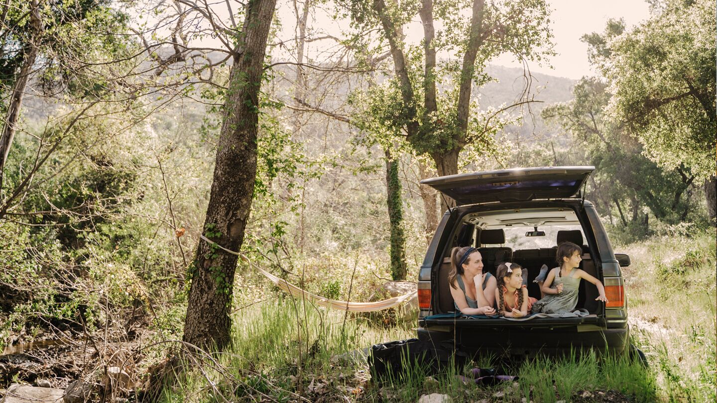 En familj sitter i bakluckan på en bil parkerad i en grön skogsmiljö med en hängmatta uppspänd mellan träden.