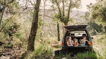 En familj sitter i bakluckan på en bil parkerad i en grön skogsmiljö med en hängmatta uppspänd mellan träden.