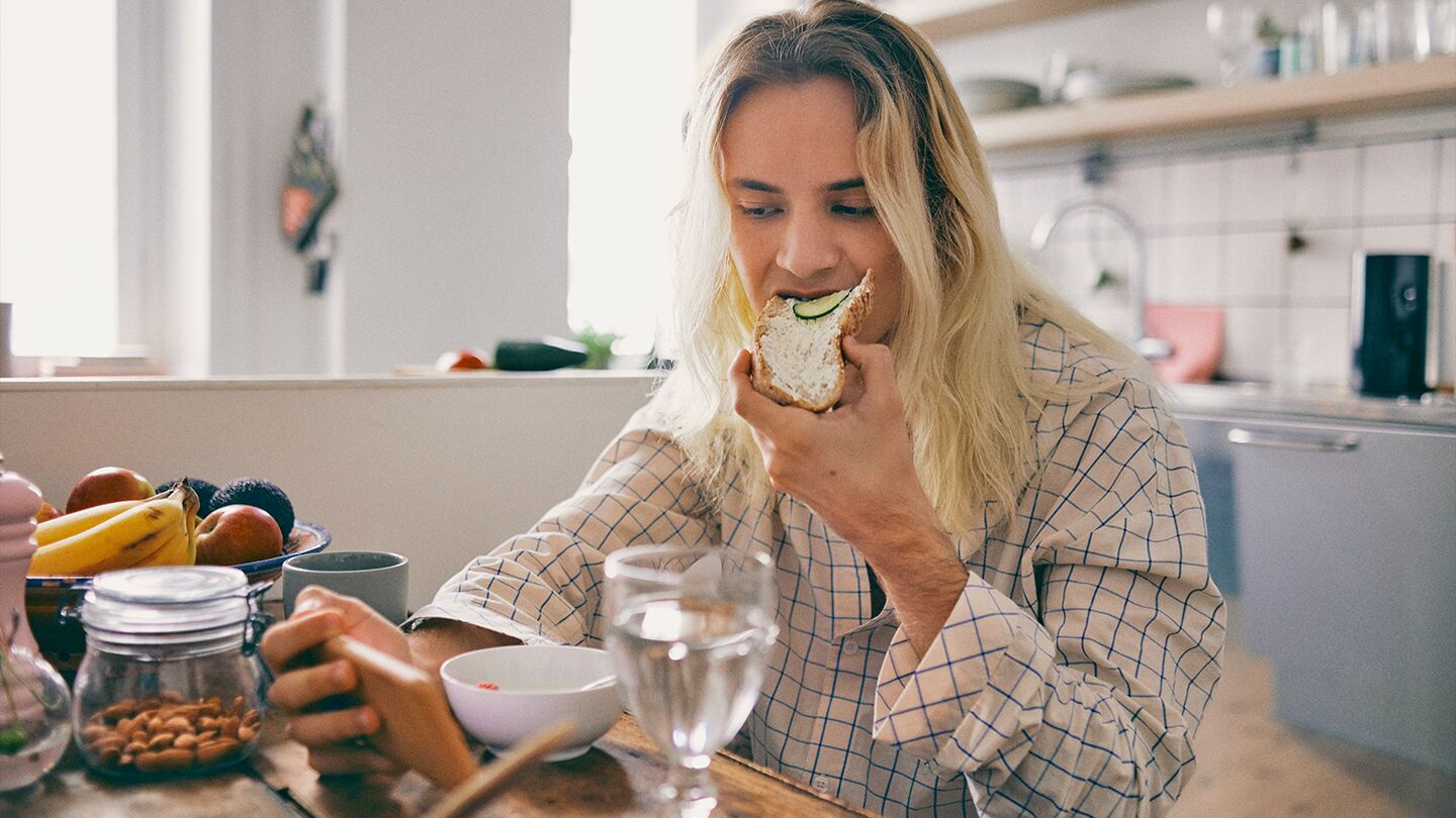 Person sitter vid ett köksbord och äter frukost med mobilen i handen.