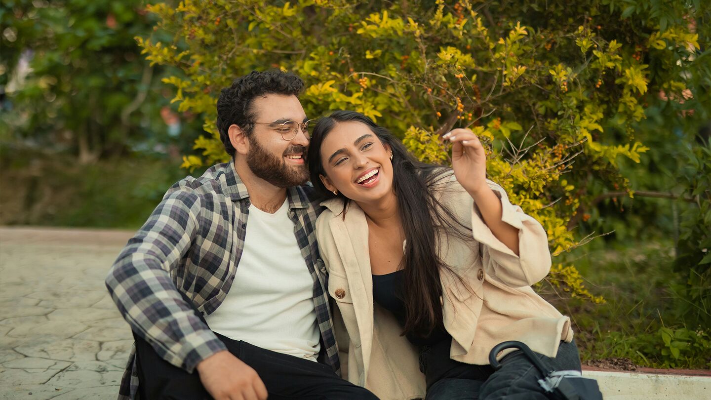 Två personer sitter tillsammans utomhus på en stenlagd yta, omgivna av grönska, och verkar njuta av en lugn stund.