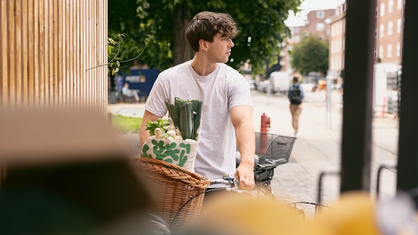 En person med en vit t-shirt som håller i en cykel med en korg fylld med gröna grönsaker.