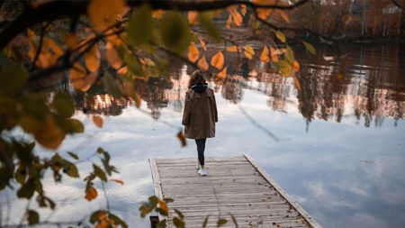 En person står på en brygga vid en stilla sjö omgiven av höstfärgade träd och löv.