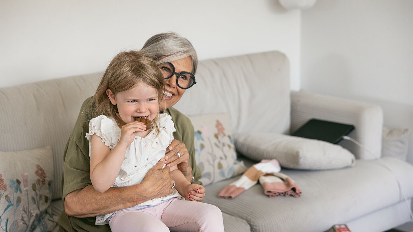 En äldre kvinna håller en liten flicka som sitter på en beige soffa med leksaker i närheten.