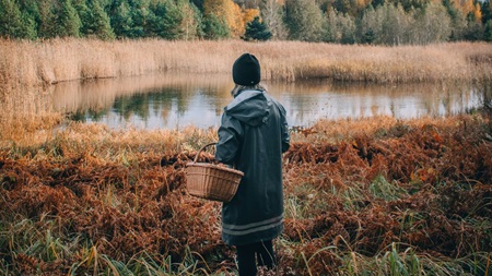 En person klädd i en mörk jacka och mössa står vid ett vatten omgivet av höstfärgad natur, med en korg i handen.