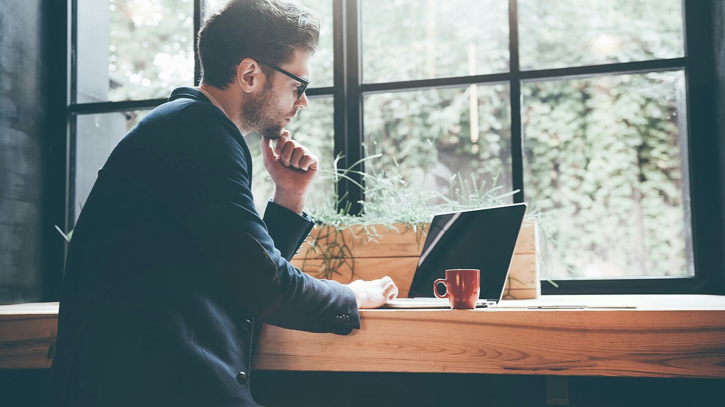 En person sitter vid ett bord med en bärbar dator och en kopp, med stora fönster och grönska i bakgrunden.