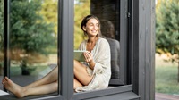 En person sitter vid ett öppet fönster med en surfplatta, omgiven av en lugn och grönskande utomhusmiljö.