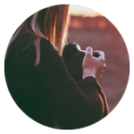 En person sitter på en trädstam med en kamera i handen, bredvid en hund som tittar ut över ett landskap i solnedgången.