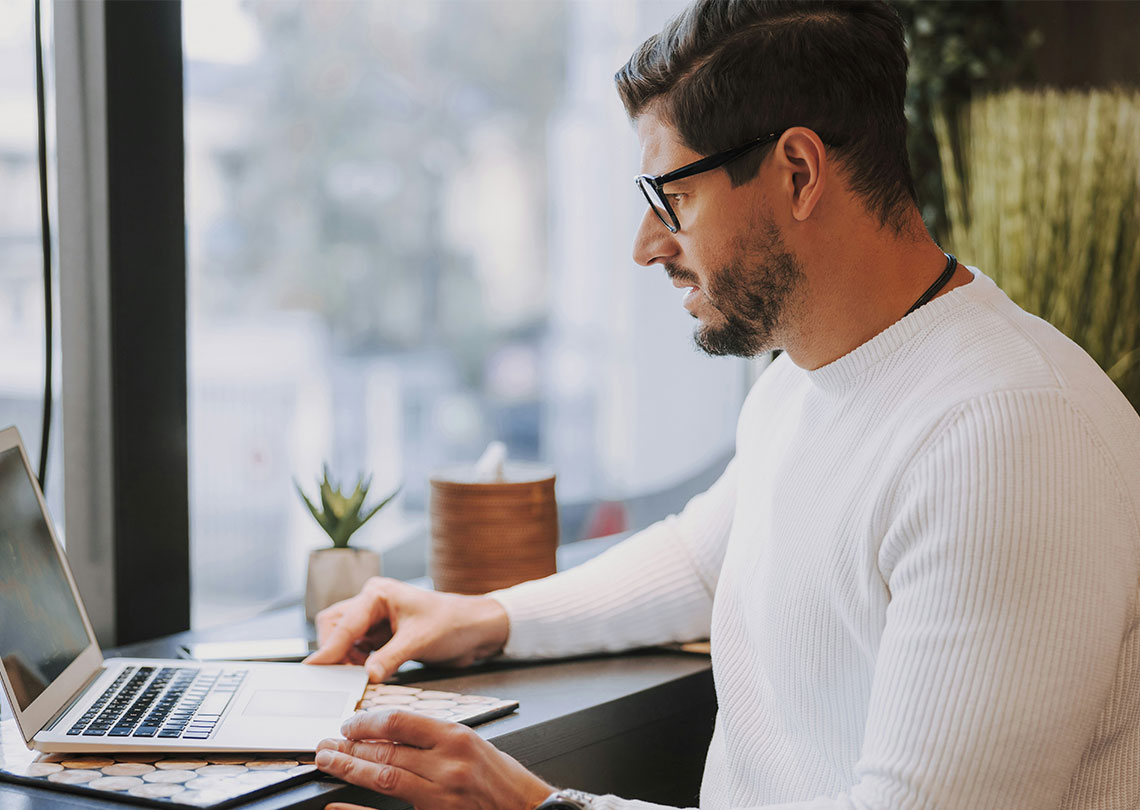 En mörkhårig man med glasögon och vit tröja arbetar på en laptop bredvid ett fönster