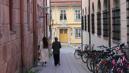 Två personer går i en smal gränd med cyklar längs ena sidan och ett gult hus i bakgrunden.