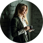 En person står inomhus vid en grå tegelvägg, håller en kopp kaffe och en fat. En grön växt syns till vänster i bilden.