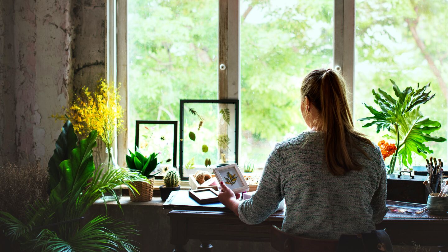 En person sitter vid ett skrivbord i ett rum med växter och tittar ut genom ett stort fönster där gröna träd syns. På skrivbordet finns papper, böcker och dekorativa föremål. Rummet har en lugn och kreativ atmosfär.