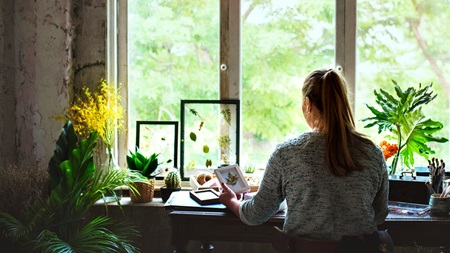 En person sitter vid ett skrivbord i ett rum med växter och tittar ut genom ett stort fönster där gröna träd syns. På skrivbordet finns papper, böcker och dekorativa föremål. Rummet har en lugn och kreativ atmosfär.