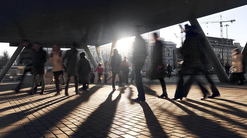 Människor promenerar i stadsmiljö med ljus och skuggor på marken.