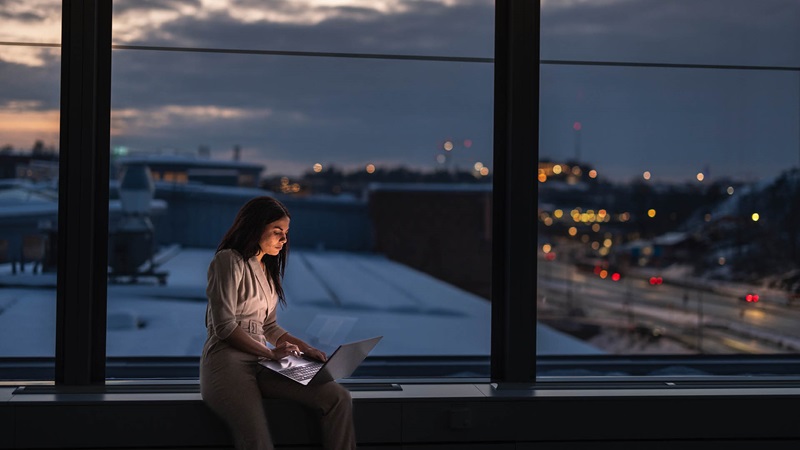 Person arbetar på en laptop vid ett fönster med utsikt över en stad i skymning.