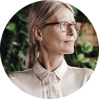 Person med ljust hår och glasögon, klädd i en ljus skjorta, sitter framför en bakgrund med gröna växter.