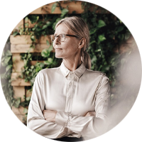 En person med blont uppsatt hår, klädd i en ljus skjorta, står med armarna i kors framför en bakgrund av gröna växter och träpanel.