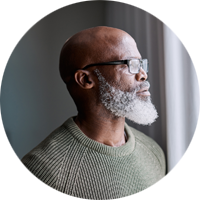 En person med rakat huvud och vitt skägg som bär en grön stickad tröja står vid ett fönster med vita gardiner och tittar ut. Ljuset från fönstret lyser upp bakgrunden.