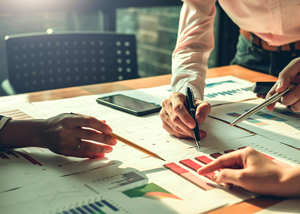 Personer som analyserar diagram och grafer på papper vid ett bord, med pennor och en mobiltelefon i närheten.