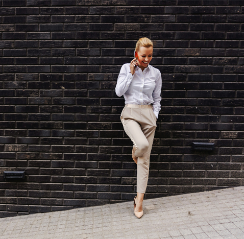 En person står framför en mörk tegelvägg och pratar i mobil, klädd i ljus skjorta och ljusa byxor.