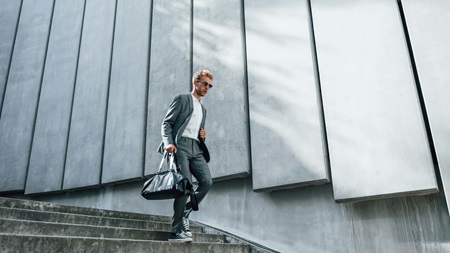 En person i grå kostym och vita sneakers går uppför en trappa, bärandes en mörk väska, framför en modern byggnad med betongpaneler.