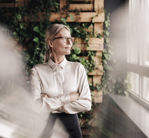 En person står vid ett fönster med grön växtvägg i bakgrunden, klädd i vit skjorta.