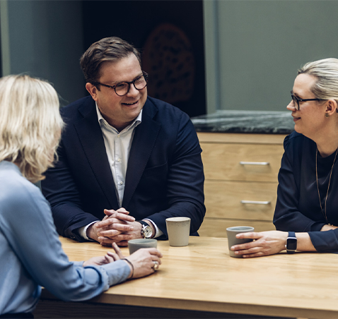  En grupp på tre personer sitter vid ett träbord och samtalar, med kaffekoppar framför sig