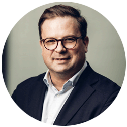 En person klädd i en mörk kostym och en ljus skjorta, fotograferad mot en grå bakgrund.