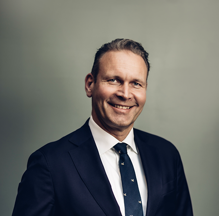 En person klädd i en mörk kostym, vit skjorta och en mörk slips med ett mönster står mot en enfärgad bakgrund.