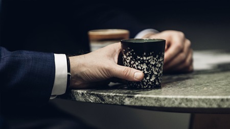 En person i kostym håller en mugg med ett svartvitt mönster vid ett bord av sten.