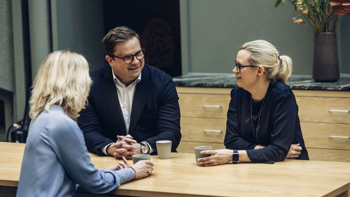 Tre personer samtalar vid ett träbord med koppar och en grön vägg i bakgrunden.